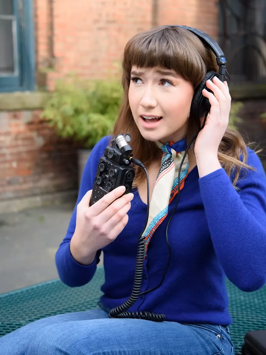 Person sitting outdoors, wearing headphones, holding an audio recorder, and looking upward with a thoughtful expression.