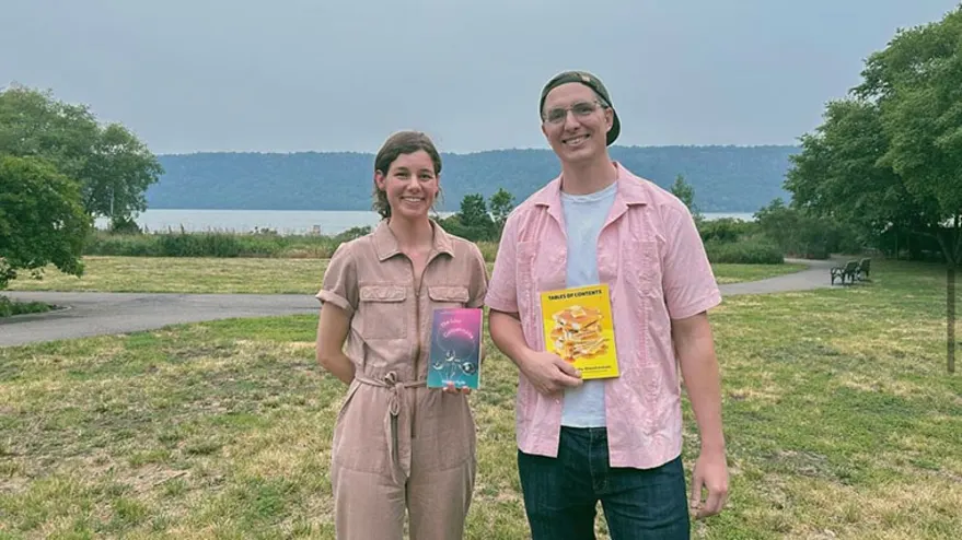 Two people stand outdoors holding books, with greenery and a river in the background.