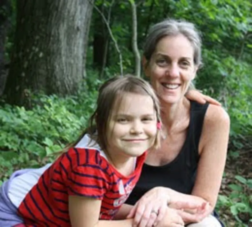 Two people smiling in a wooded area, one with an arm around the other.
