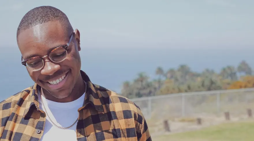 Person smiling, wearing glasses and a checkered shirt, standing outdoors with a blurred background of trees and ocean.