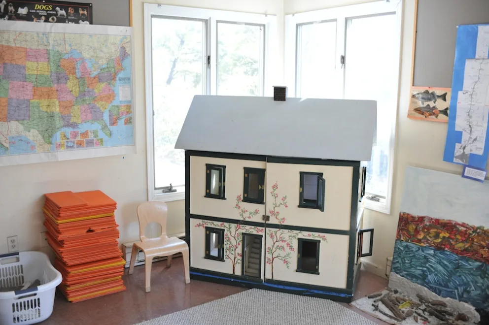Large dollhouse in a room with maps on the walls and a chair nearby.