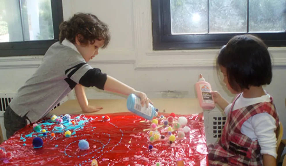 Two children engaged in a craft activity with paint and pom-poms on a table.