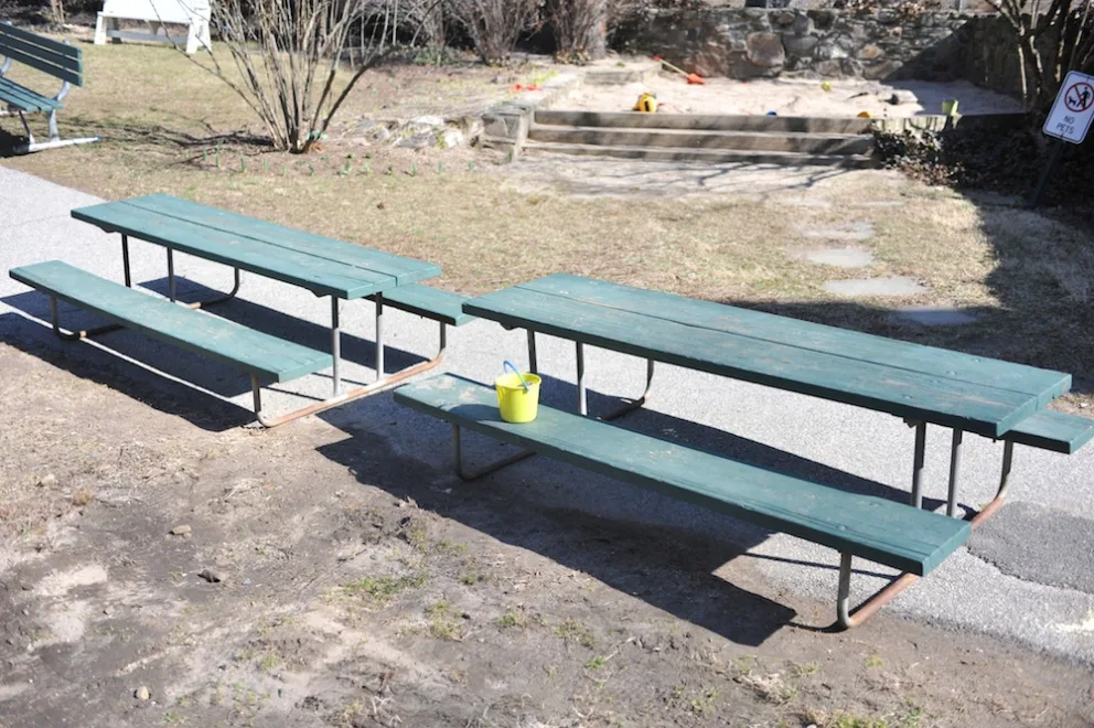 Two green picnic tables with a yellow bucket on one, set in a yard.