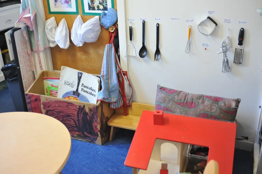 Classroom corner with play kitchen, utensils on wall, books, and cushioned seating.