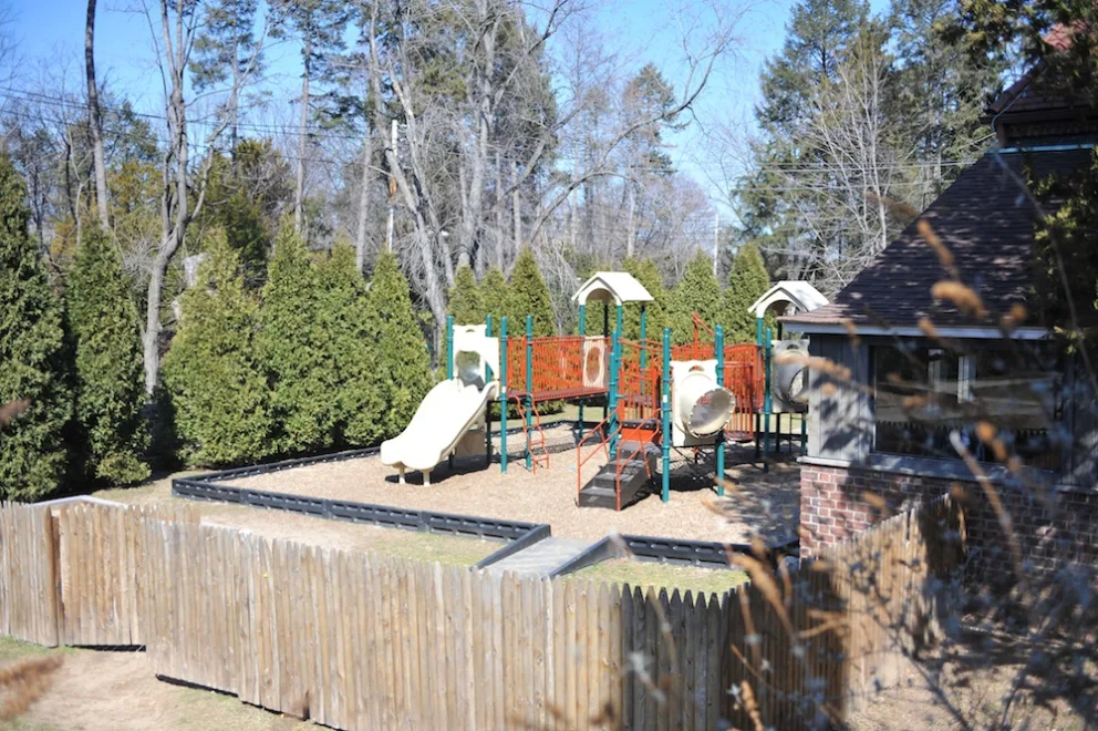 Playground with slides and climbing structures, surrounded by trees and a wooden fence.