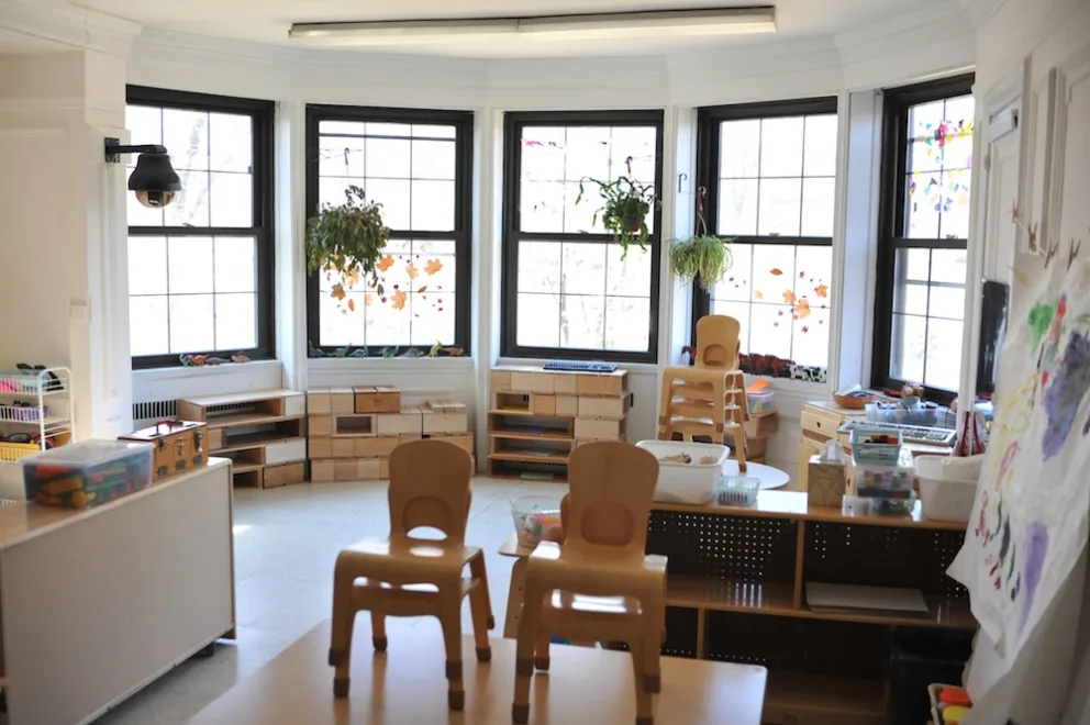 Sunny classroom with child-sized chairs, plants by windows, and art supplies.