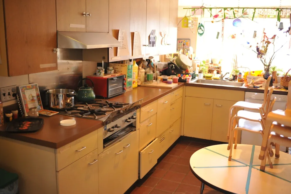 A cluttered kitchen with yellow cabinets and a stove, with natural light coming in.