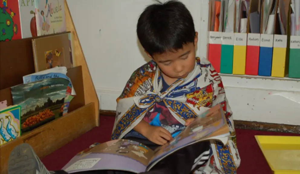 Child reading a book in a library corner with bookshelves nearby.