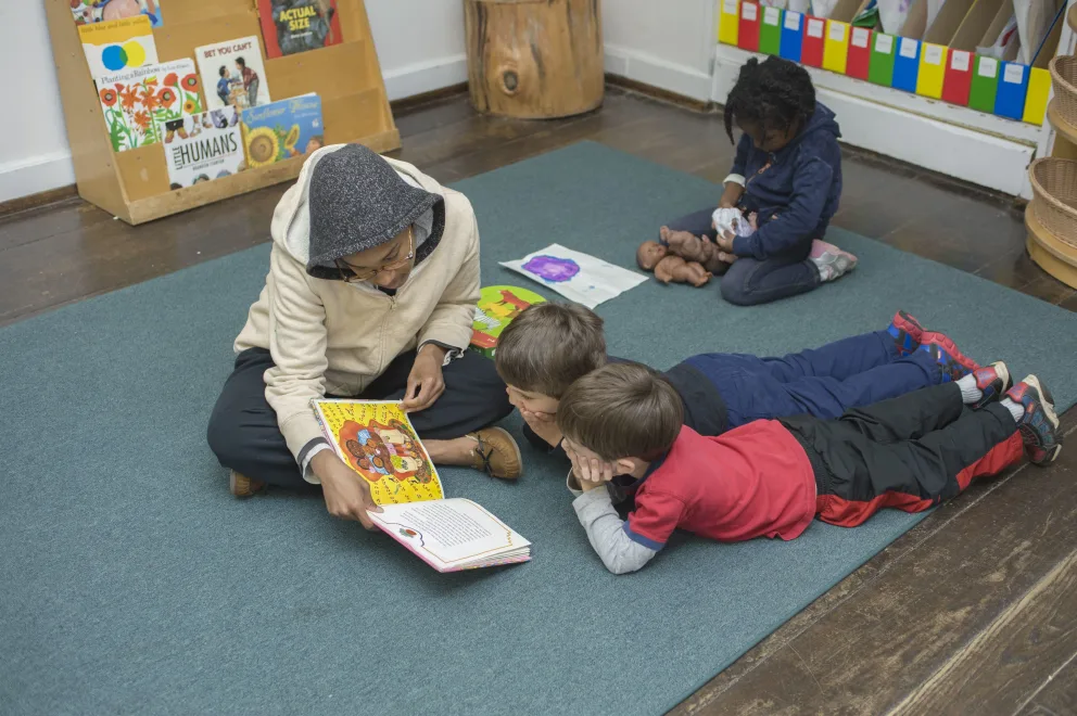 Adult reading a book to children on a floor mat in a room with toys.