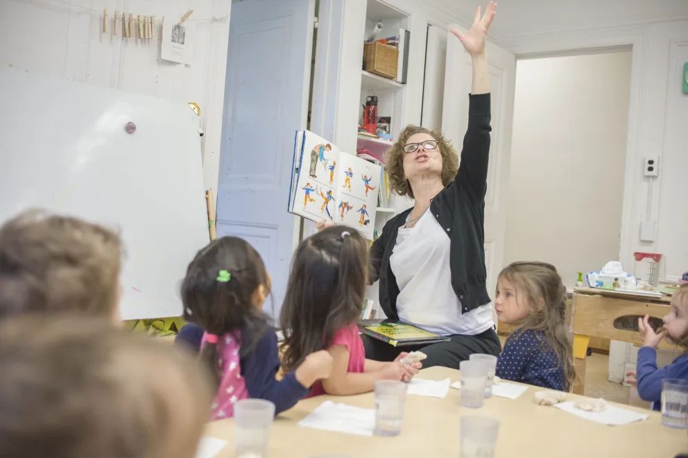 Teacher reading to children with arm raised in a classroom setting.