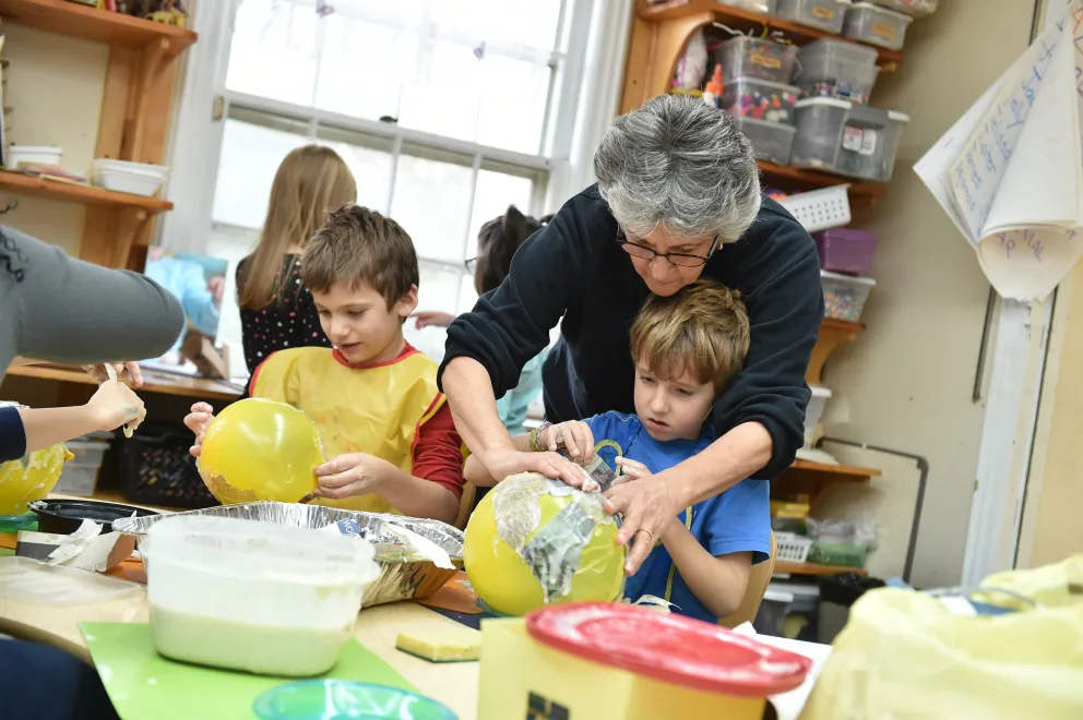 Adult assisting child with a craft project in a classroom setting.