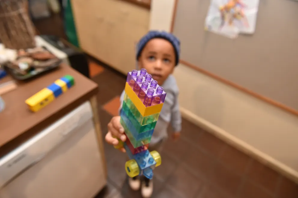 Child in a knit hat proudly displaying a colorful block tower indoors.