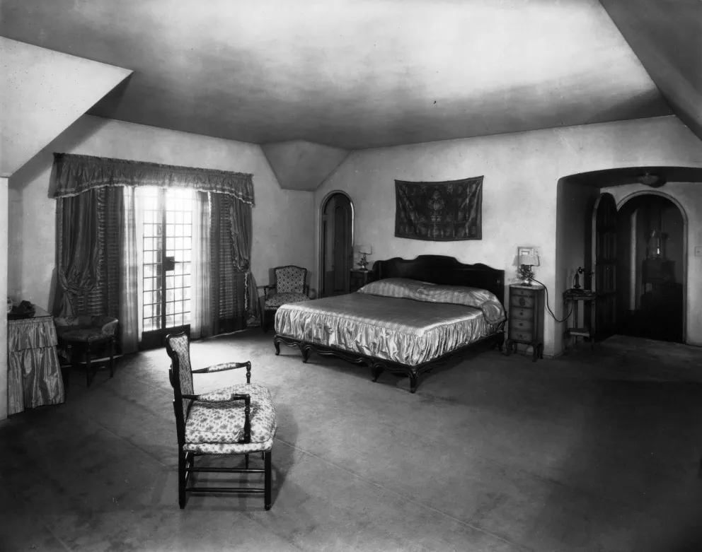 Vintage bedroom with large bed, chair, patterned curtains, and arched doorway.