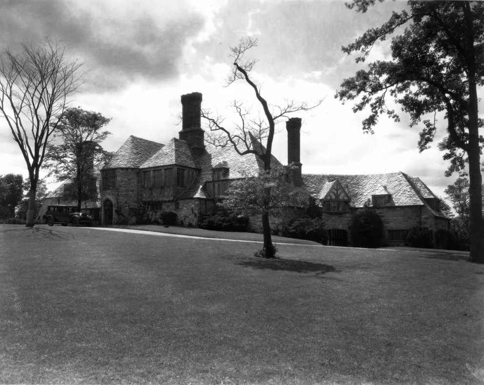 Large stone house with tall chimneys surrounded by trees and lawn.