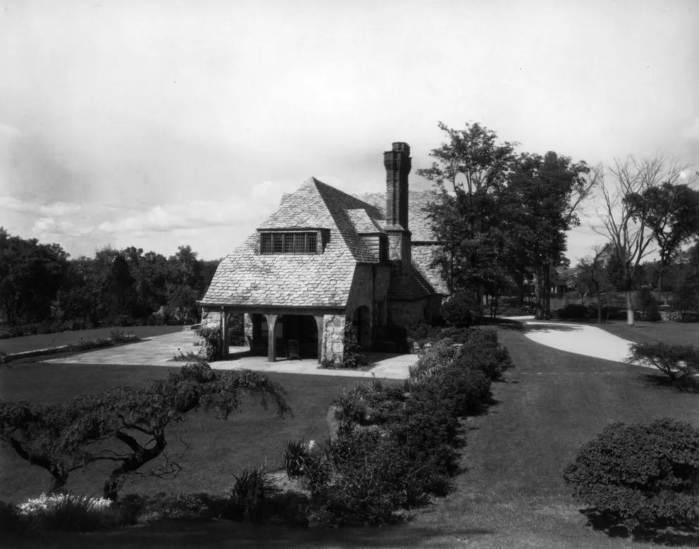 Vintage photo of a stone cottage with a large chimney, surrounded by manicured gardens.