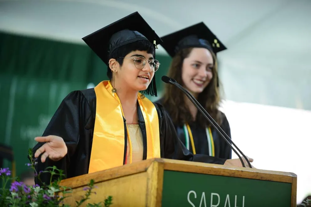 Individual giving a speech at graduation with another person listening beside them.