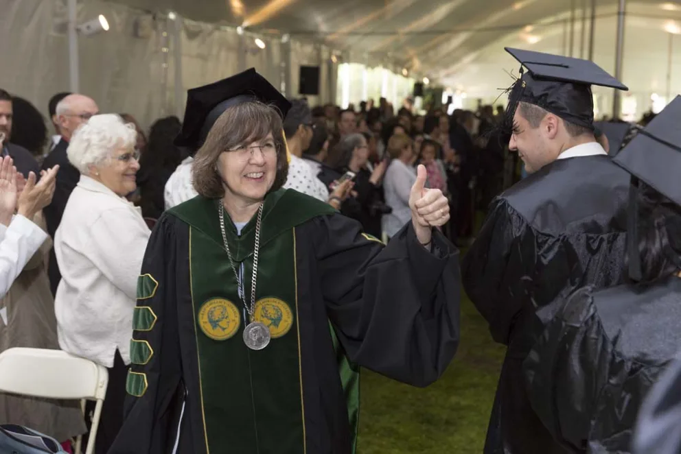 Individual in academic regalia giving a thumbs-up at graduation ceremony.