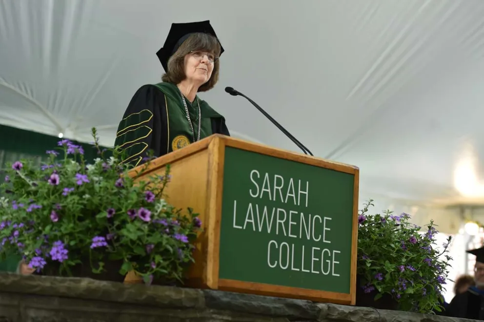 Individual in academic regalia speaking at a podium with a college name.