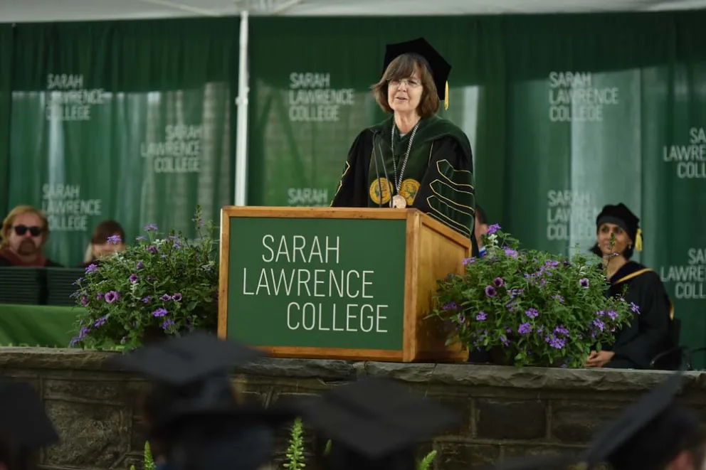 Person in academic regalia speaking at a podium marked 'Sarah Lawrence College' with attendees in caps and gowns.
