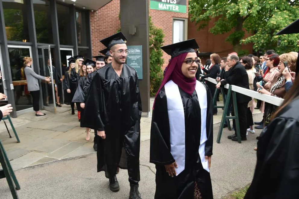 Graduates in caps and gowns walking, with onlookers nearby.