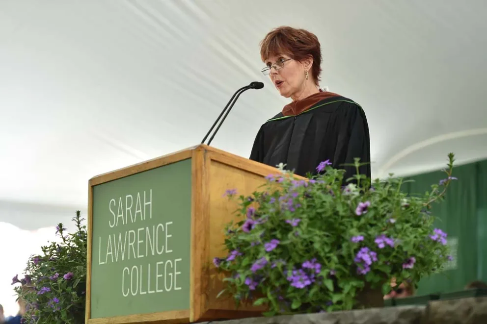 Individual speaking at a podium with "Sarah Lawrence College" visible, at an event with floral decorations.