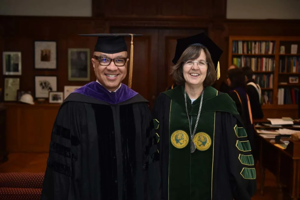 Two individuals in academic regalia smiling in a room with bookshelves.