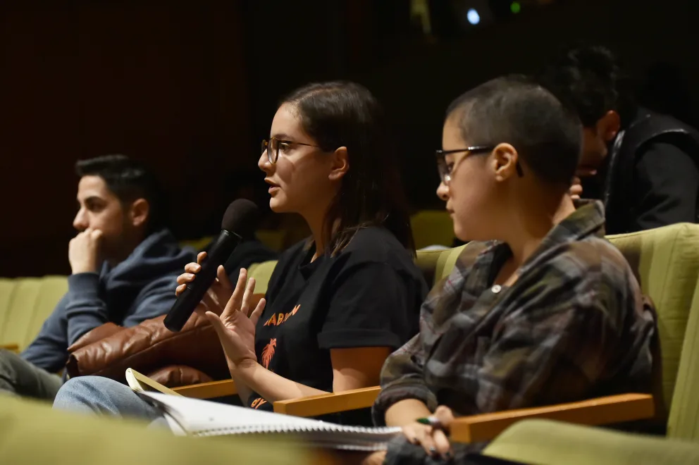 A person is speaking into a microphone during a panel discussion while sitting on a green couch with other participants who appear to be listening attentively.