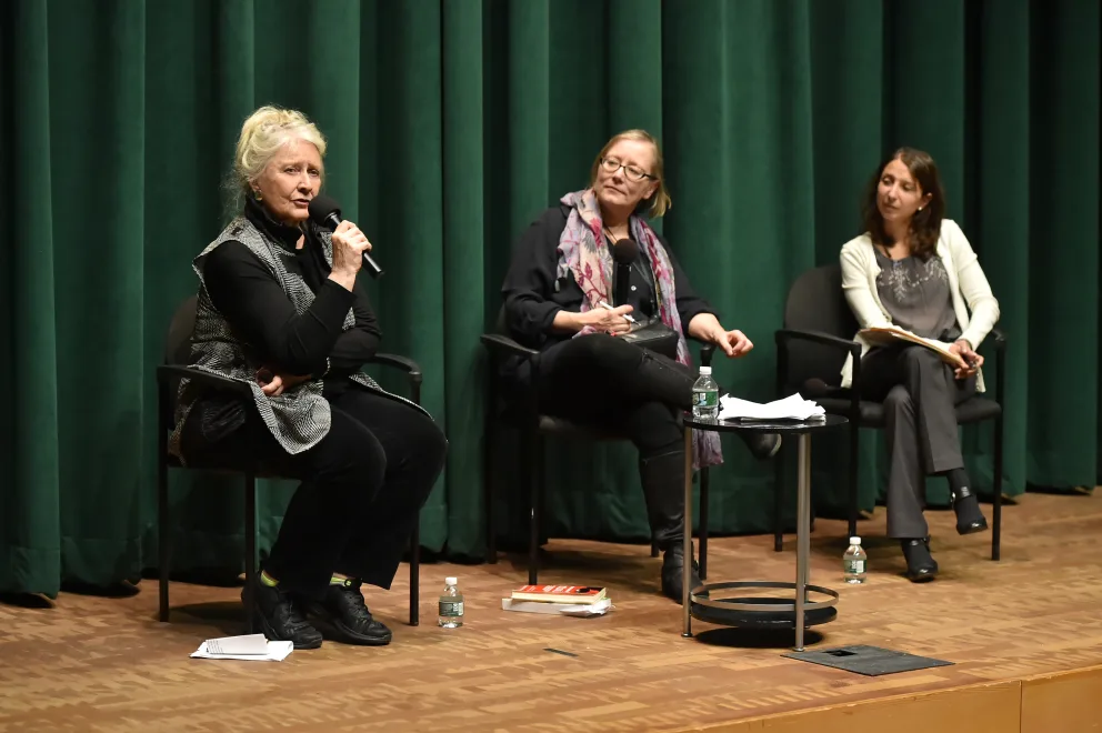 Three people sitting on stage, one speaking into a microphone, with papers and water bottles on a small table, against a green curtain background.