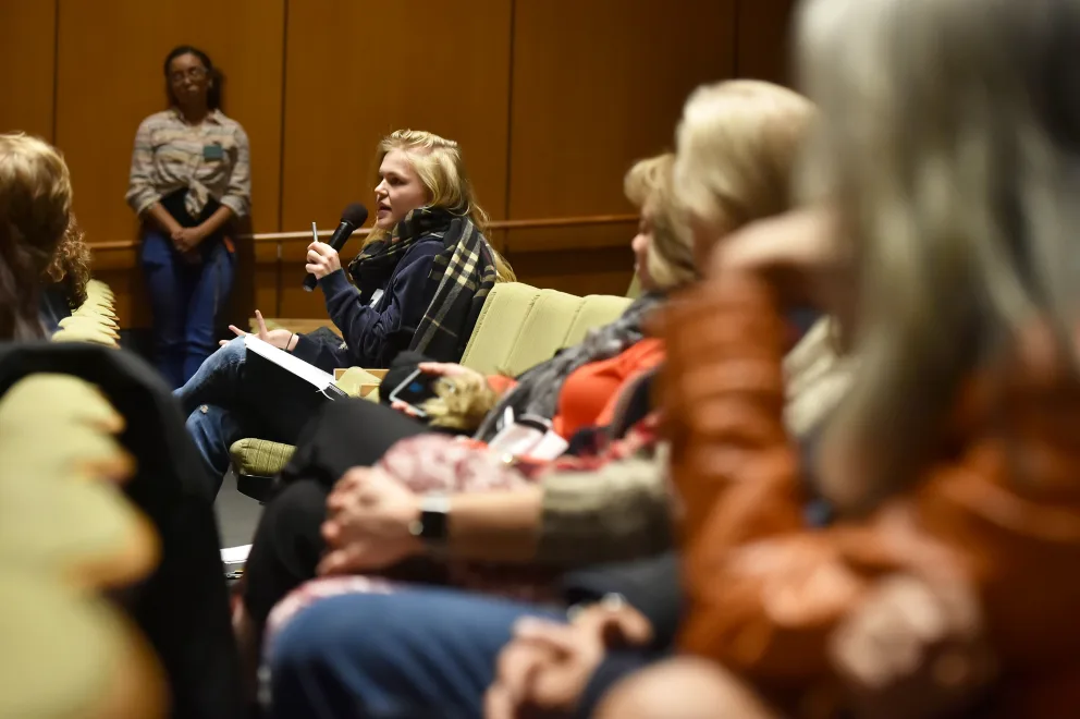A woman sitting and speaking into a microphone during a discussion with an audience in a conference room setting. Other participants are listening attentively.
