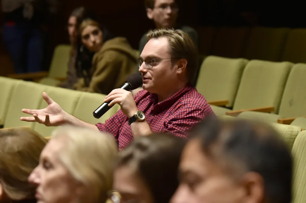A person in a red checkered shirt speaking into a microphone in an auditorium with other attendees in the background.