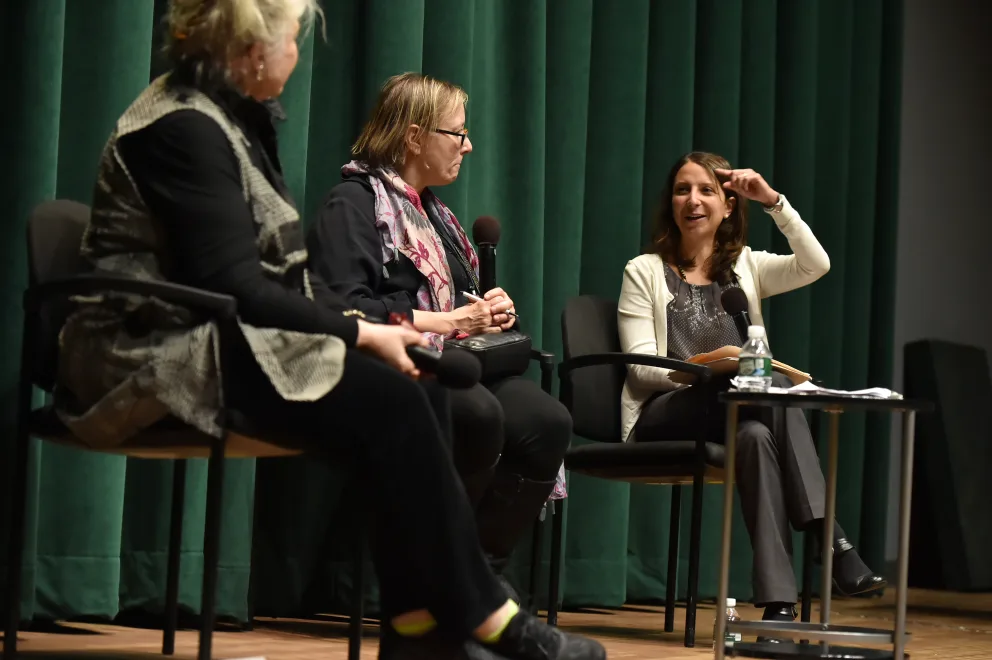 Three people sitting on stage in a panel discussion, with one person gesturing while speaking to an audience. The setting appears to be an event or conference with a green backdrop.