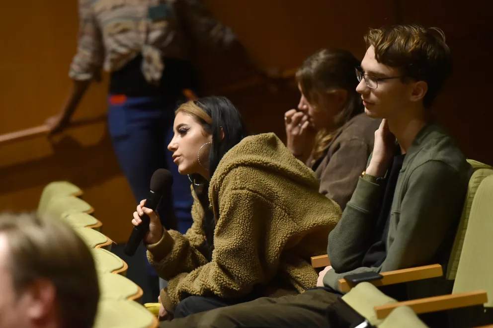 A person holding a microphone seated in a lecture hall, speaking or preparing to speak, surrounded by other attendees.