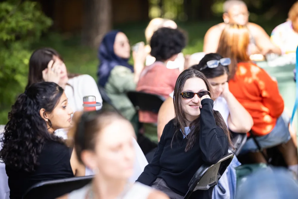 People sitting at tables outdoors, socializing and enjoying a sunny day. A person in sunglasses is smiling, with others conversing around them.