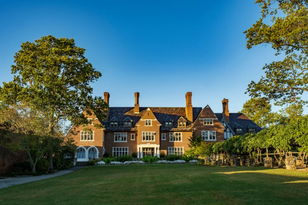 Large brick mansion with multiple chimneys and gabled roofs, surrounded by green trees and lawn under a clear blue sky.