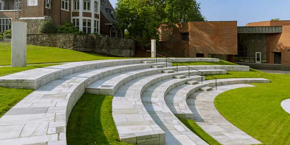 Curved stone amphitheater surrounded by green grass, with brick and stone buildings in the background.