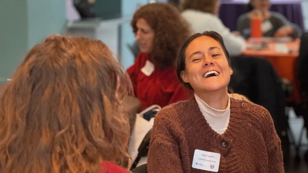 Person wearing a name tag is laughing in a group setting indoors. Others are seated in the background, focused on conversations.