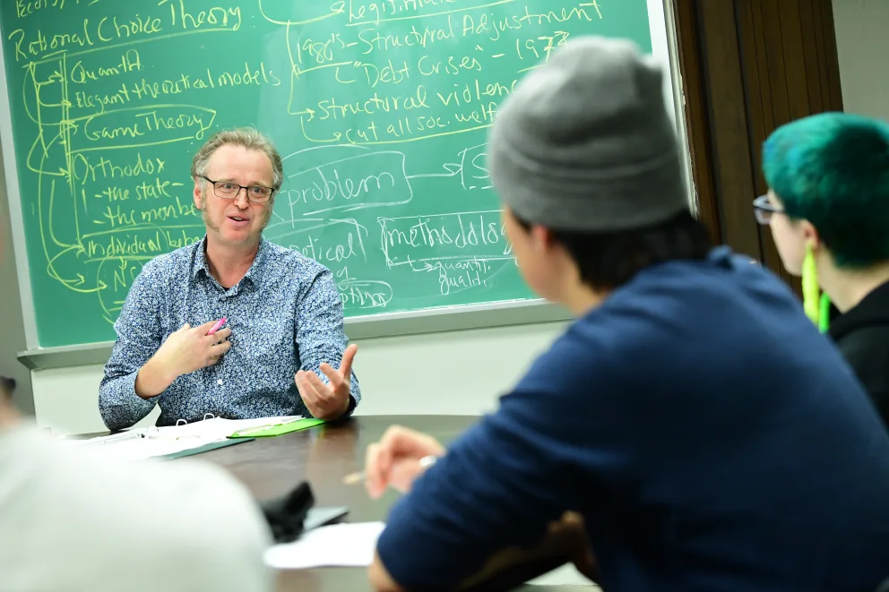 Person gestures while speaking to two others at a table in a classroom. Chalkboard in the background with complex notes.