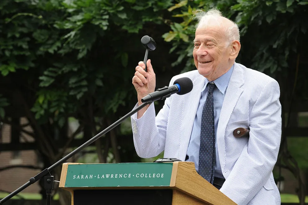 Person in a light suit holds a gavel, speaking at a podium with "Sarah Lawrence College" sign, greenery in the background.