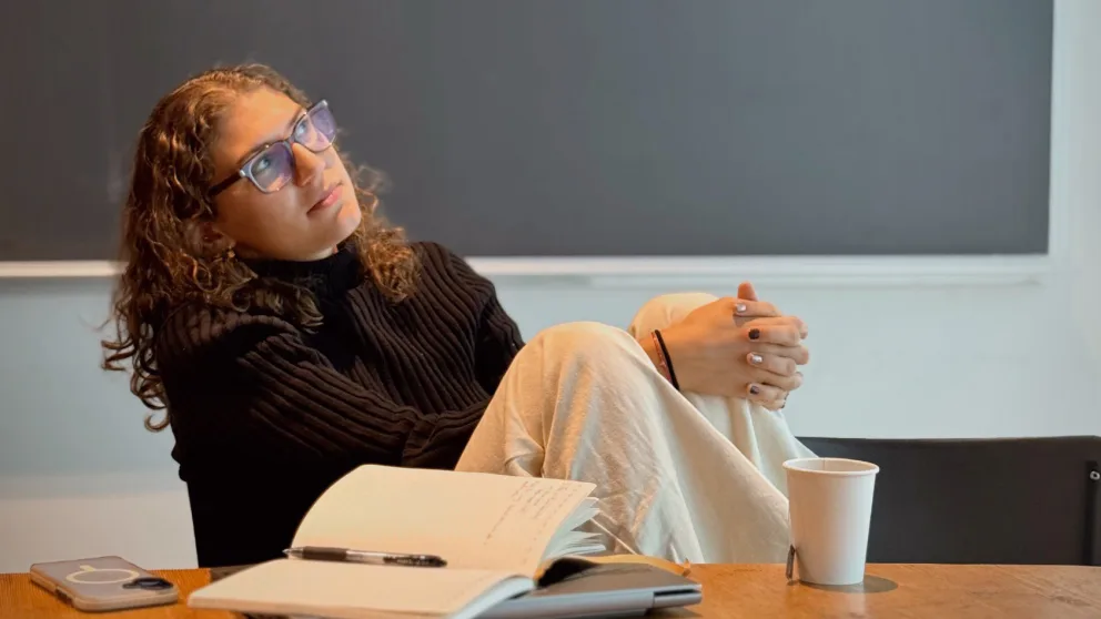 Person with curly hair and glasses sits at a table with a notebook, pencil, phone, and paper cup, gazing contemplatively to the side.