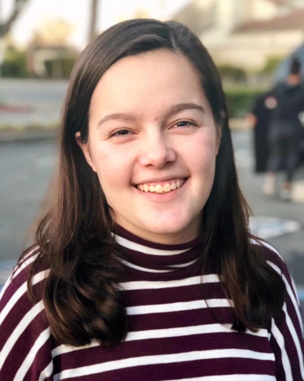 Person smiling outdoors, wearing a maroon and white striped shirt. Blurred background with trees and buildings.
