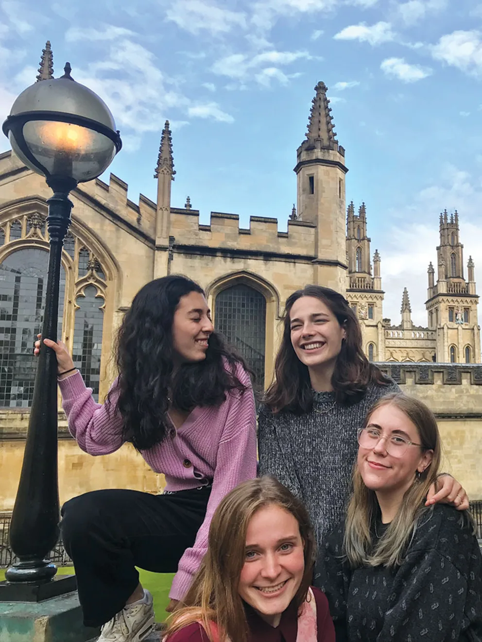 Four people smiling, posing near a historical building with spires, under a bright sky and next to a streetlamp.
