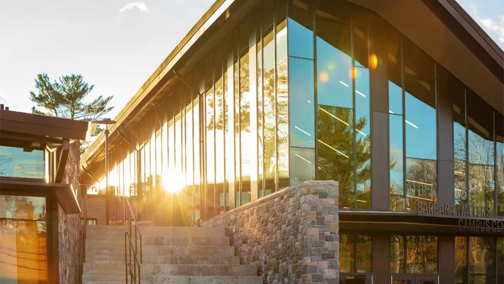 Modern building with glass walls reflecting sunlight, surrounded by stonework and trees. Steps lead to an entrance on the right side.