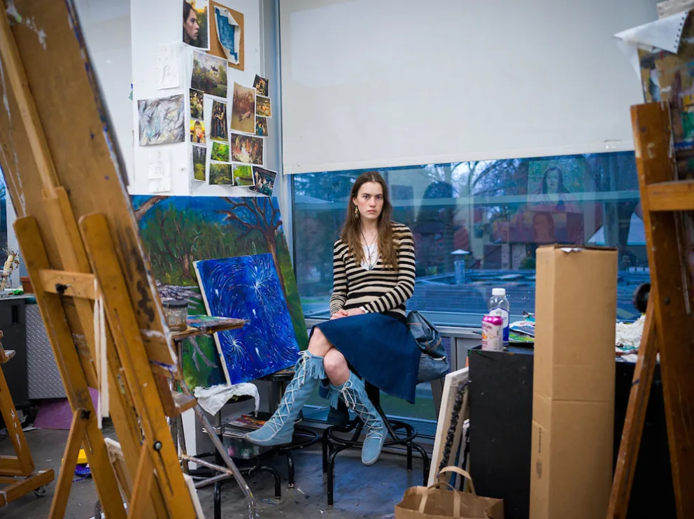 Person sitting in an art studio with paintings, easels, and art supplies around. Large window in the background.