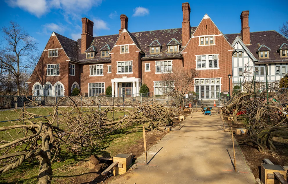 Large brick building with multiple chimneys and gabled roofs, set behind a path lined with trimmed trees and wooden benches on a sunny day.