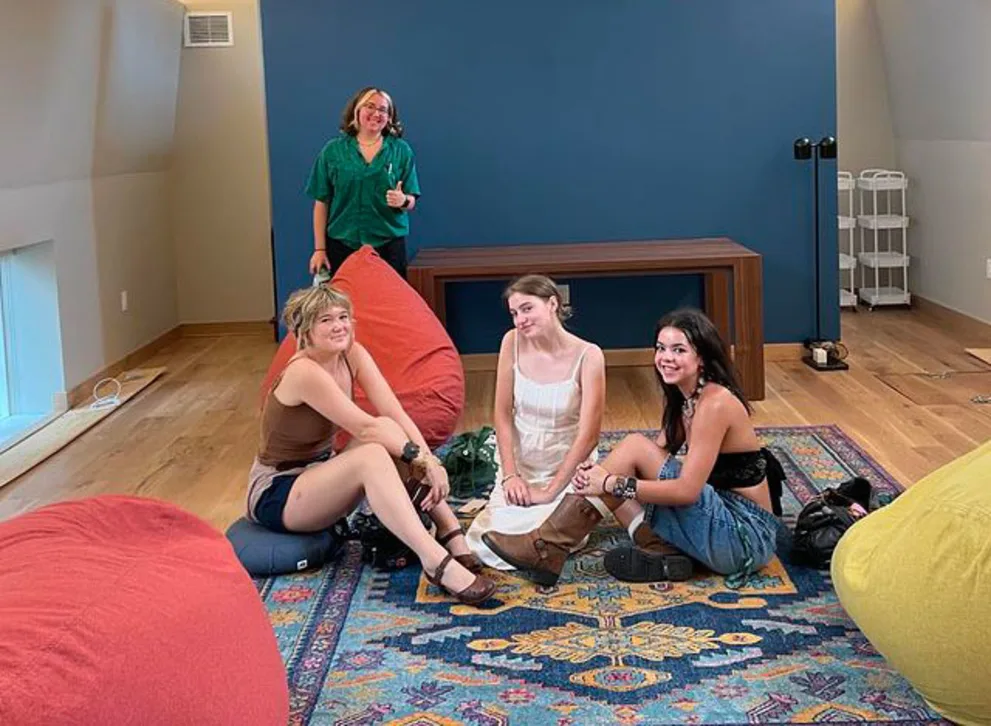 Three people sit on a colorful rug surrounded by bean bags, one person stands in the background. Wooden floor, blue wall, and a table in the room.