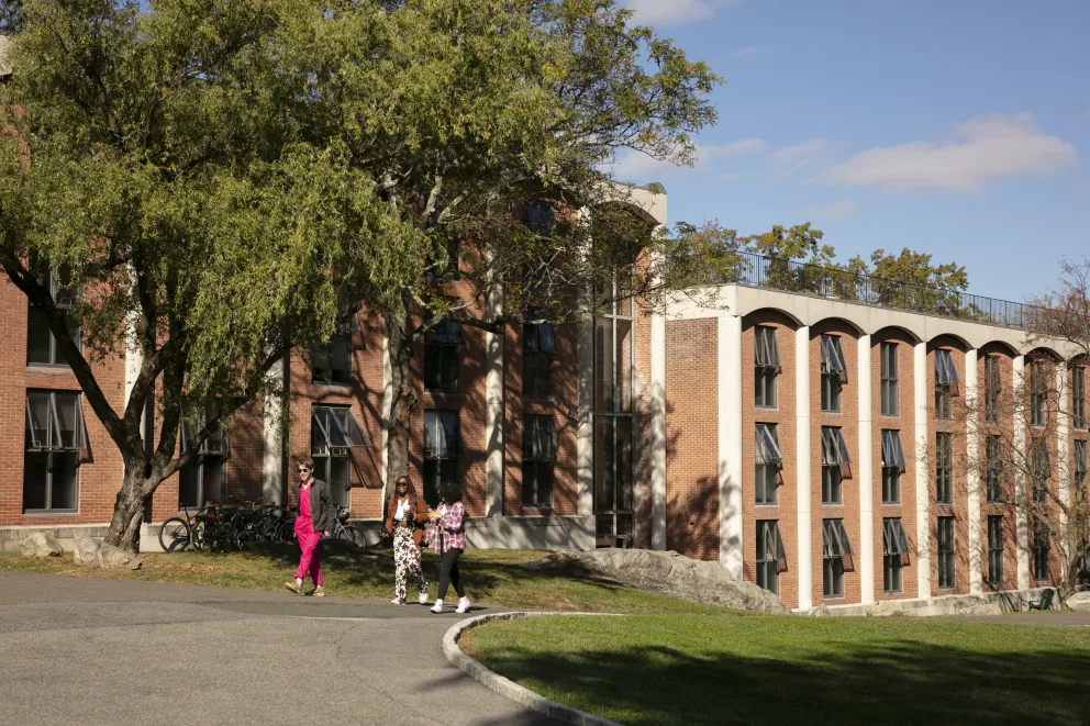 Three people walk on a path near a large brick building with many windows, surrounded by trees on a sunny day.