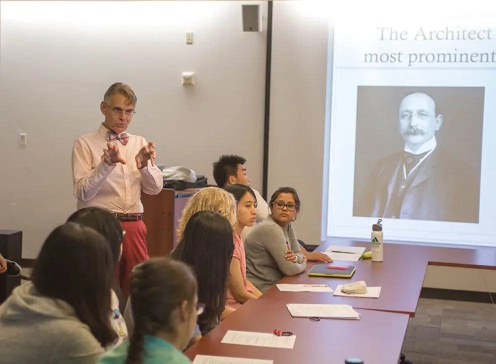 Person in pink shirt gestures while speaking to a group seated at a table, with a historical figure's image projected on the screen.