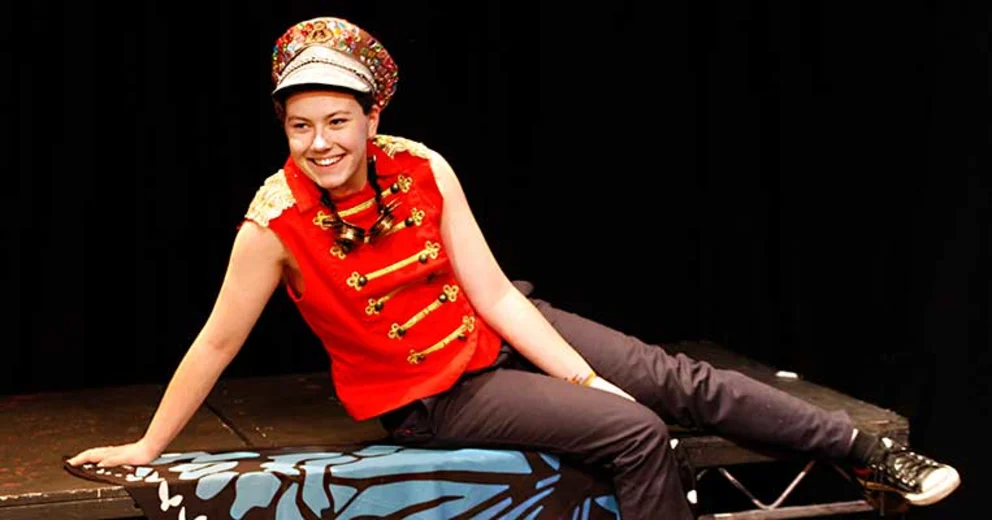 Person in a colorful military-style hat and red vest lounges on a stage prop with a butterfly design, smiling under stage lights.