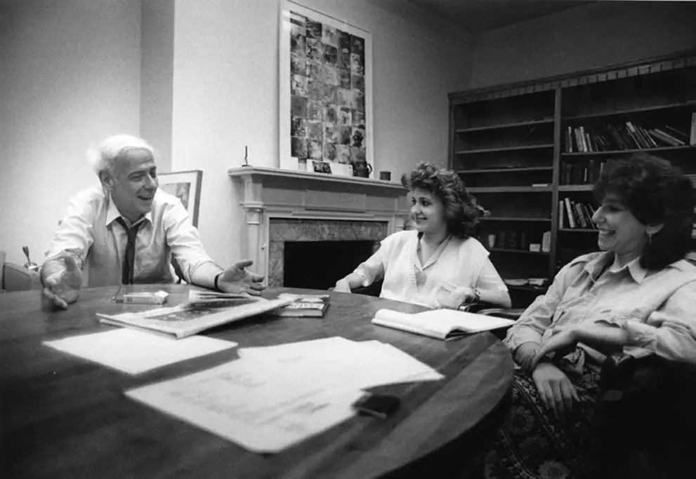Three people sitting at a table in a room with bookshelves, discussing and smiling. One person is gesturing with hands.