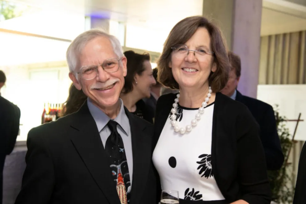 Two people smiling at a formal event, surrounded by others in the background. One holds a glass and wears a floral-patterned outfit.